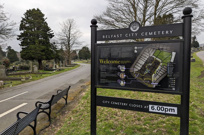 The entrance sign at Belfast City Cemetery which is black with a map of the cemetery and in the background you can see a road leading away and graves