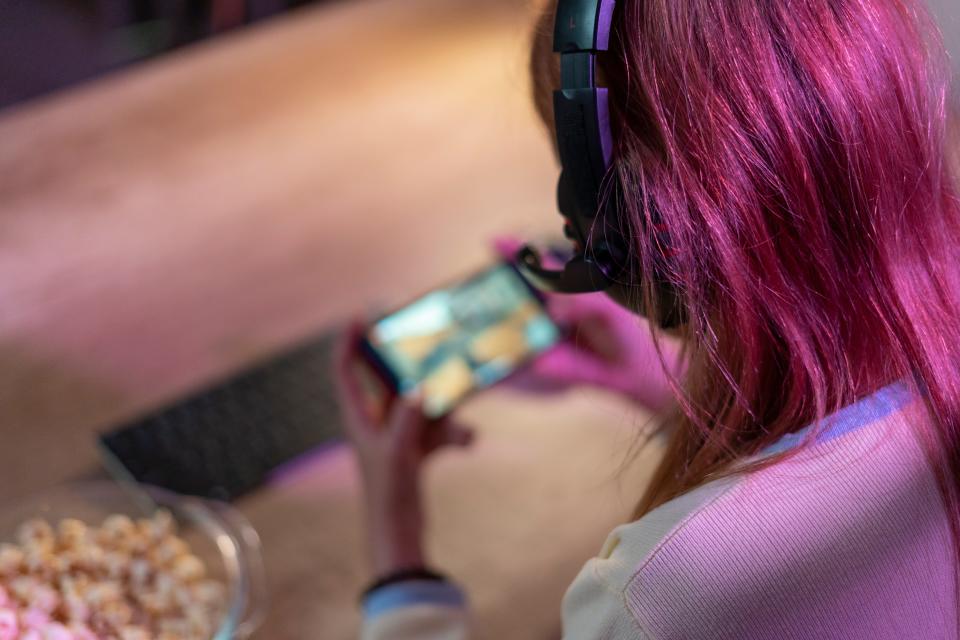 A girl with pink hair and a gaming headset sits on a wooden floor next to a bowl of snacks while playing a game on her phone.