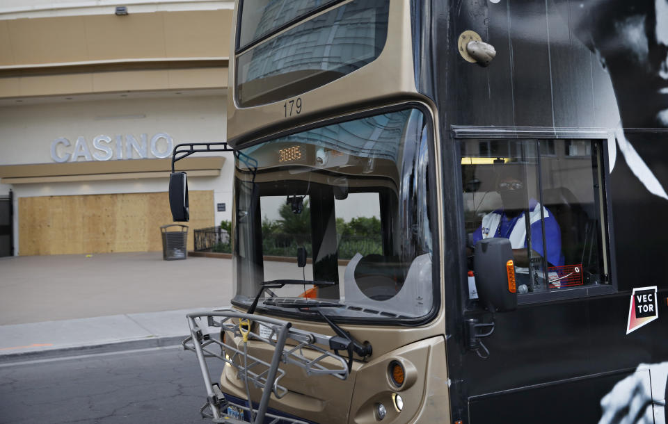 In this April 21, 2020, photo, a bus driver in a mask drives by a boarded-up casino along the Strip in Las Vegas. Bus service in parts of the city has been reduced during the coronavirus shutdown. (AP Photo/John Locher)