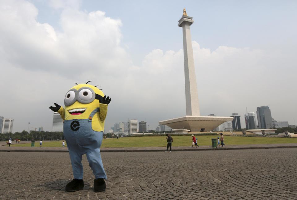 In this Thursday, May 8, 2014 photo, a street performer dressed as minion character from the movie "Despicable Me" waits for tourists to pose for a photo with him for small change, as the National Monument is seen in the background in Jakarta, Indonesia. The 132-meter (433-feet) tall monument, a popular landmark in the capital, is being cleaned for the first time in more than two decades. (AP Photo/Dita Alangkara)