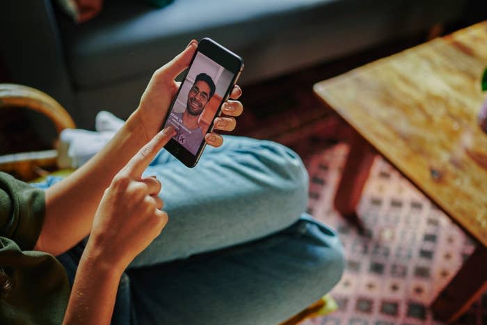 Una persona a gambe incrociate siede su un divano e tiene in mano uno smartphone che mostra l'immagine di un uomo sorridente durante una videochiamata