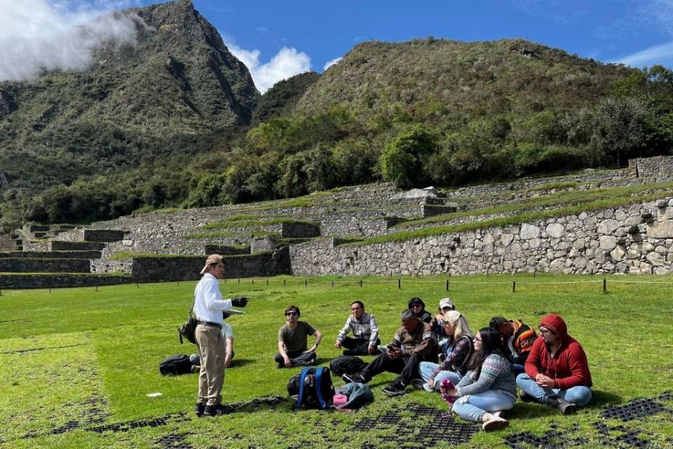 A group of tourists listen to a guide give a lecture at an ancient ruin site surrounded by mountains