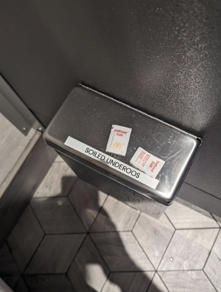 A metal disposal bin labeled 'SOILED UNDERPOOS' mounted on a wall above a tiled floor