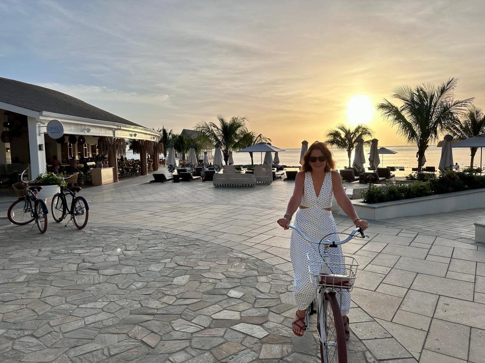 Terri Peters is riding a bike at sunset, wearing a white dress and sunglasses. She is at a resort with palm trees in the background.