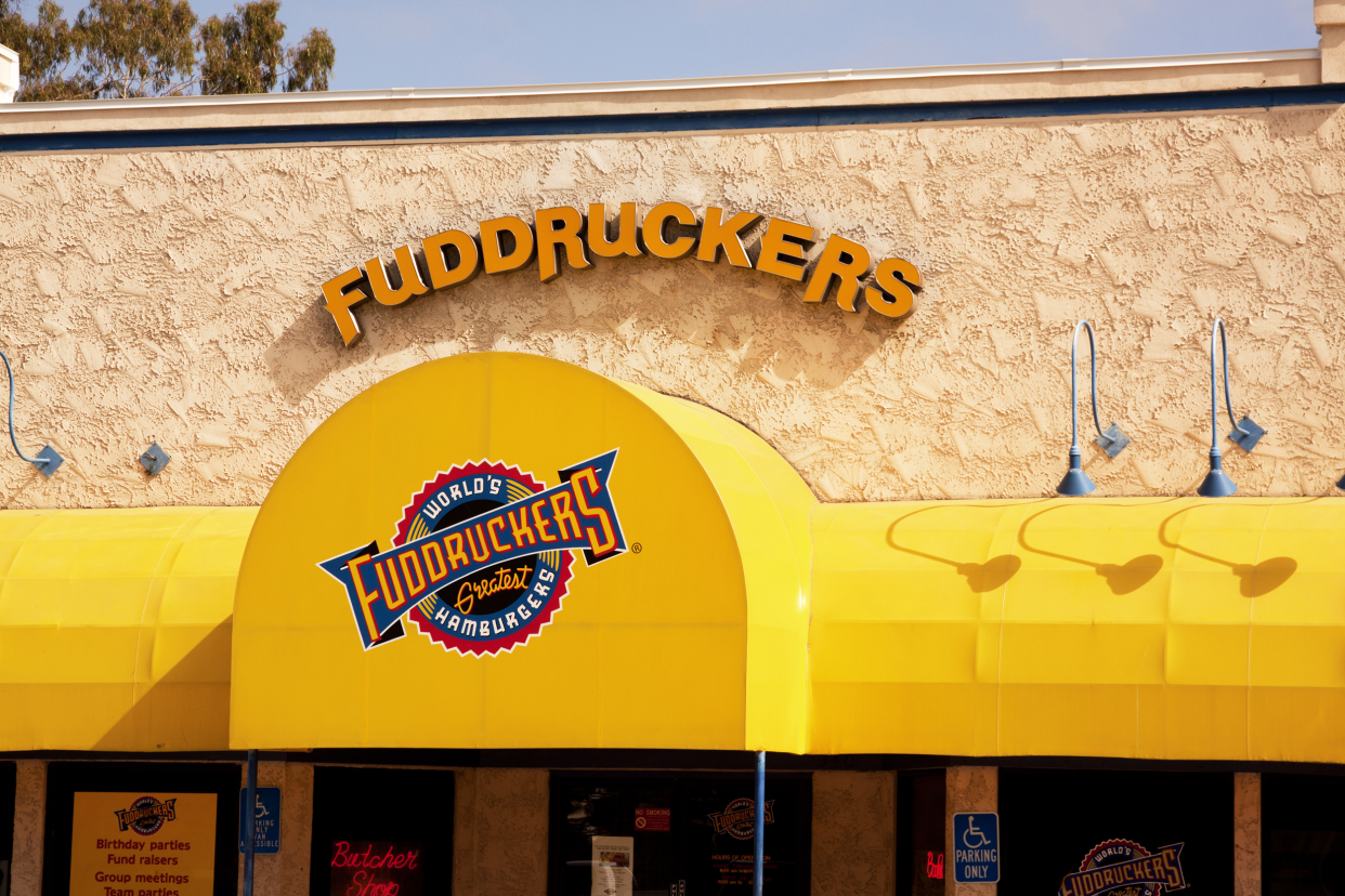 Closeup of front exterior Fuddruckers restaurant in Chula Vista, California with bright yellow porch roof against a sliver of blue sky