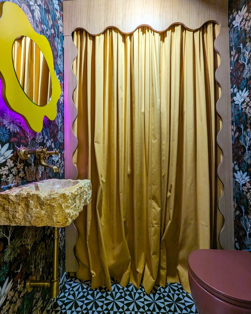 Bathroom with navy botanical wallpaper, yellow wavy mirror, and gold curtain