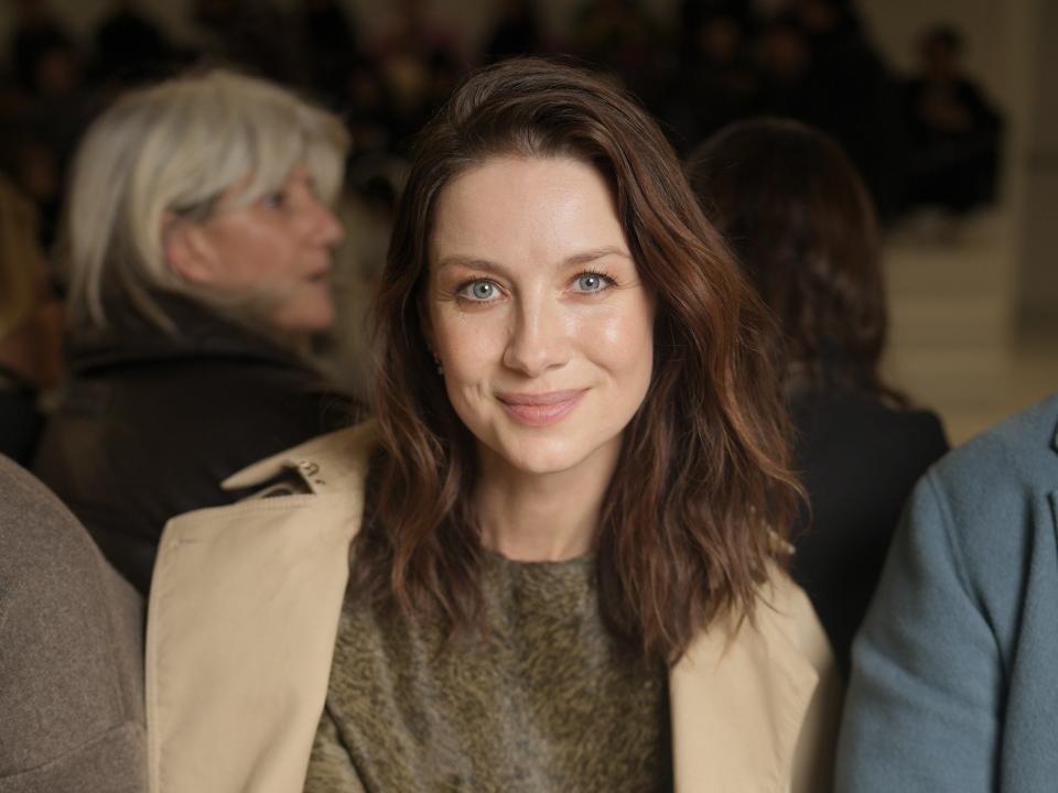 Caitríona Balfe standing in amongst a large group of people while wearing a tan coat and smiling.