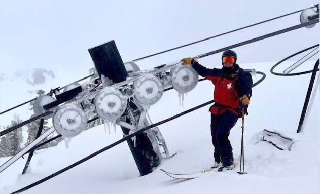 A skiier stands next to a chair lift buried in snow.