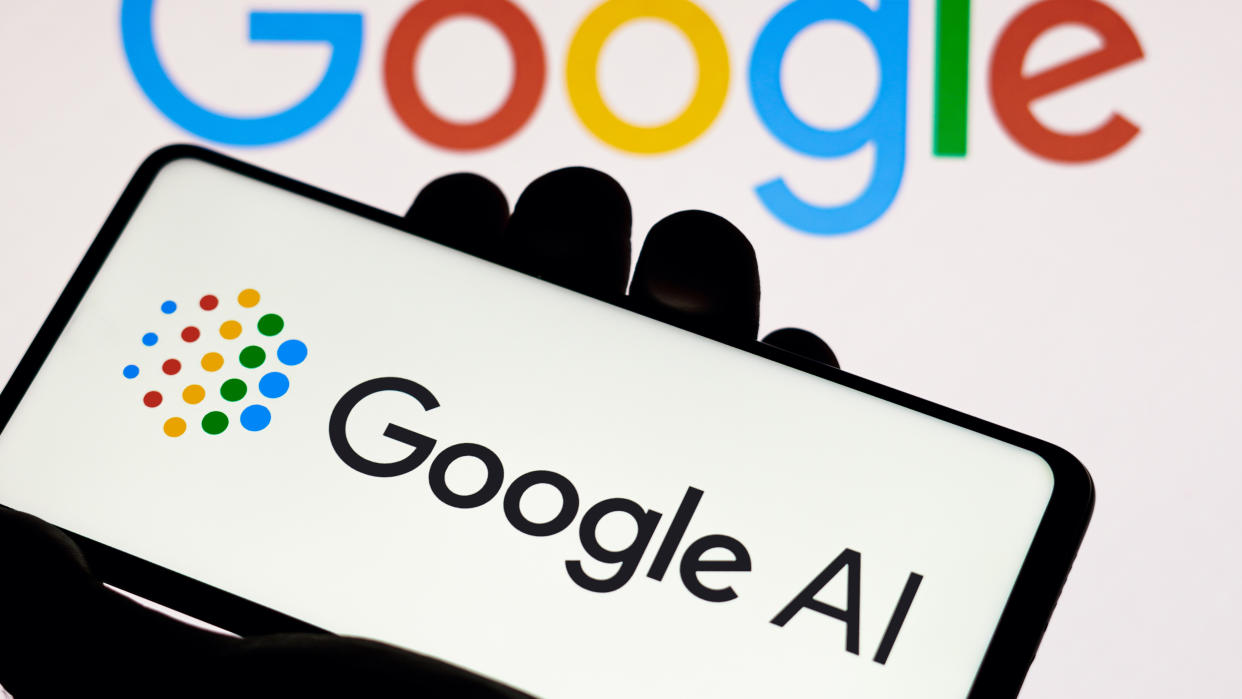  A Google AI logo is on a phone held in a hand, in front of a Google Logo. 