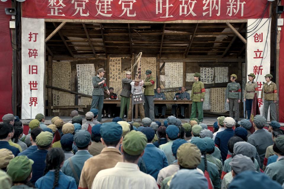 a struggle session during the cultural revolution, depicted fictionally on screen in 3 body problem. a man wearing a nameplate and large cone hat is being held with his arms behind his back in front of a microphone. students in green uniforms and red armbands stand on stage with him, as a crowd of people looks on