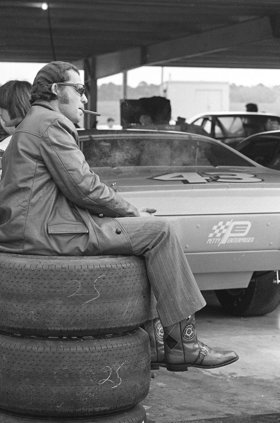 FILE -Stock car racer Richard Petty, of Randleman, N.C., sits on a stack of tires, smoking a cigar, while his crew prepares his 1972 Plymouth for a practice run before qualifying Saturday, Feb. 12, 1972, for the Daytona 500, at Daytona International Speedway in Daytona, Fla. NASCAR’s next 75 years almost certainly will include at least a partially electric vehicle turning laps at Daytona International Speedway. It’s unfathomable to some, unconscionable to others. . (AP Photo/James P. Kerlin, File)