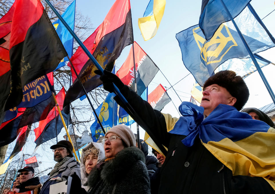 Activists wearing coat and hats, one draped in a flag, hold signs and various flags aloft.