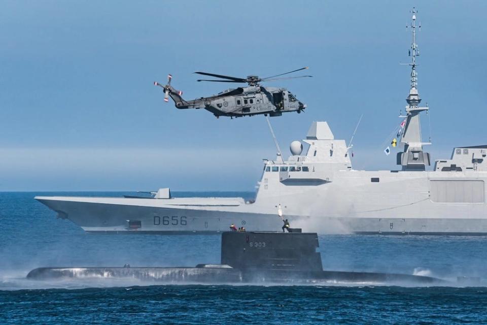 French Navy frigate FNS Alsace is seen in the background as a CH-148 Cyclone Maritime Helicopter conducts a hoist exercise