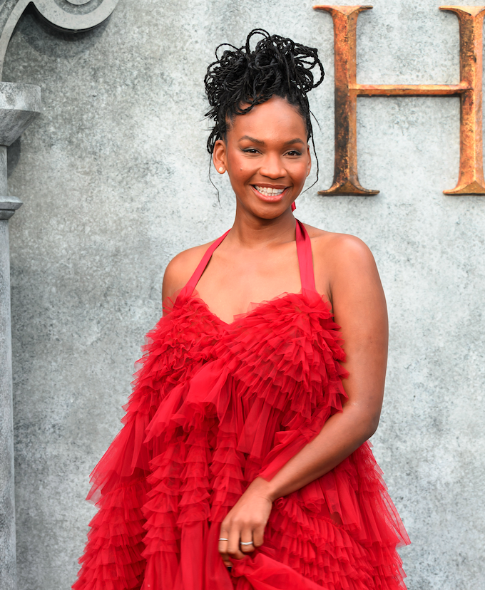 Woman in an elegant red, layered ruffle dress, smiling in front of a stone wall with a decorative "H" behind her