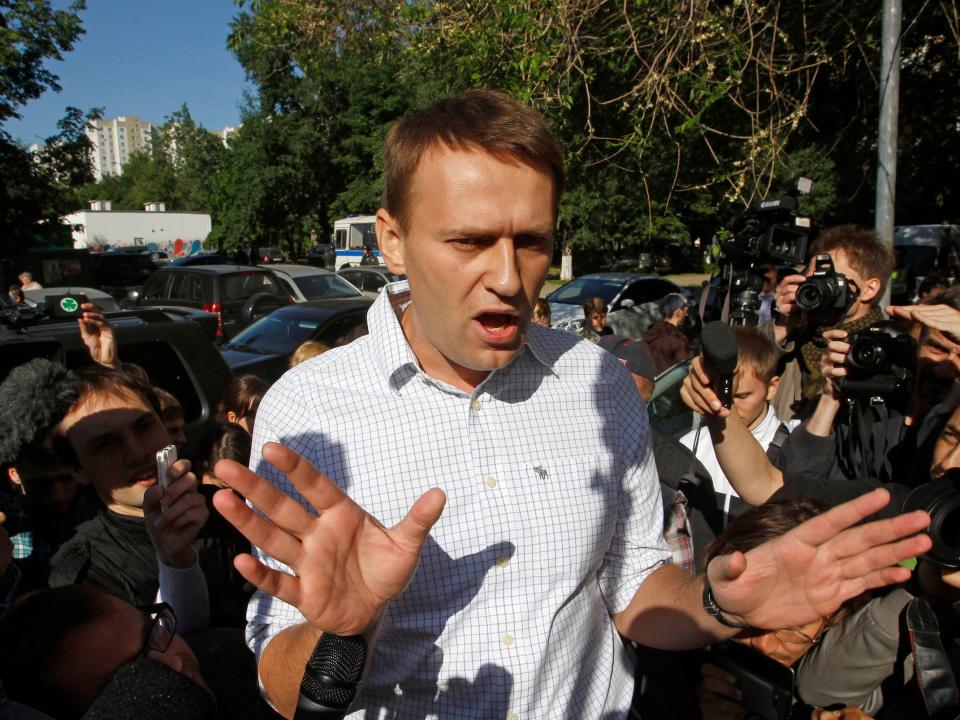 Alexei Navalny speaks to his supporters and media holding his hands in the air as members of th e press hold microphones to him and photographers point cameras at him.