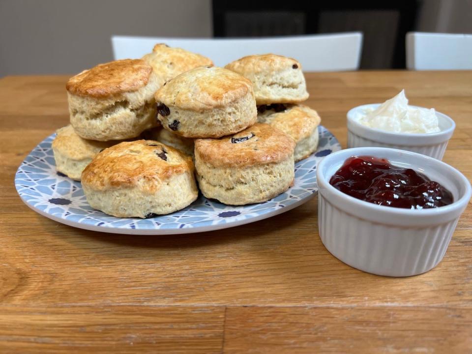 A plate with a blue and white print with scones piled on top of it, next to white ramekins containing strawberry jam and a faux clotted cream, on a wooden kitchen island.