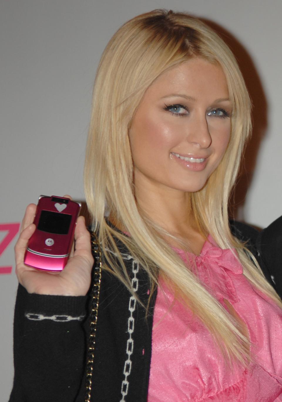 Paris Hilton holds a flip phone and smiles. She wears a pink blouse and a black cardigan with chain details