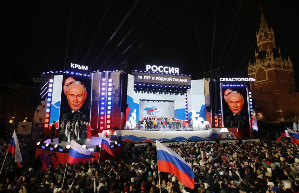 A crowd waving Russian flags in front of a stage, with Vladimir Putin pictured on large TV screens.