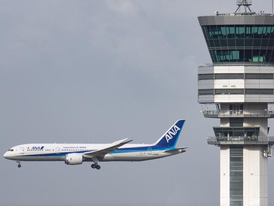 An ANA Boeing 787 Dreamliner flying past an air traffic control tower during the day.
