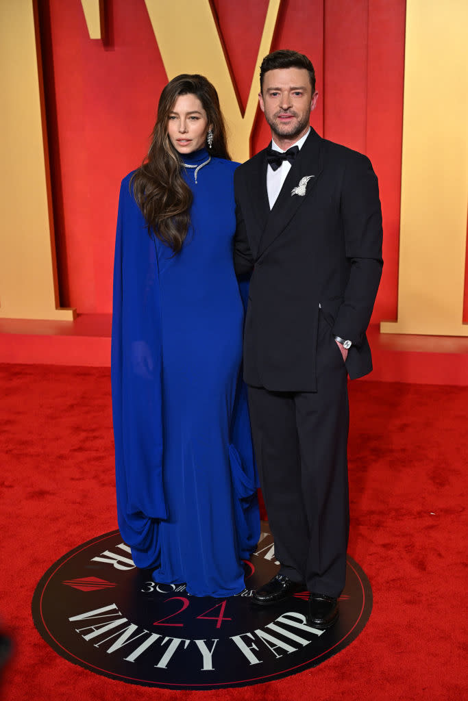 Two individuals posing, one in a blue evening gown with a cape, the other in a black suit and bow tie