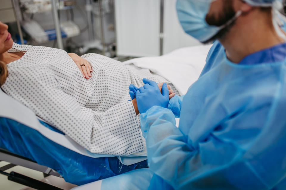 A healthcare professional wearing protective gear holds hands with a pregnant woman who is lying on a hospital bed