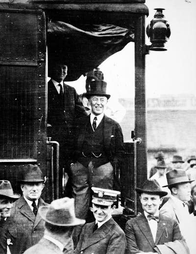 A white man wearing a dark suit and top hat stands at the rear of a train and is surrounded by onlookers.