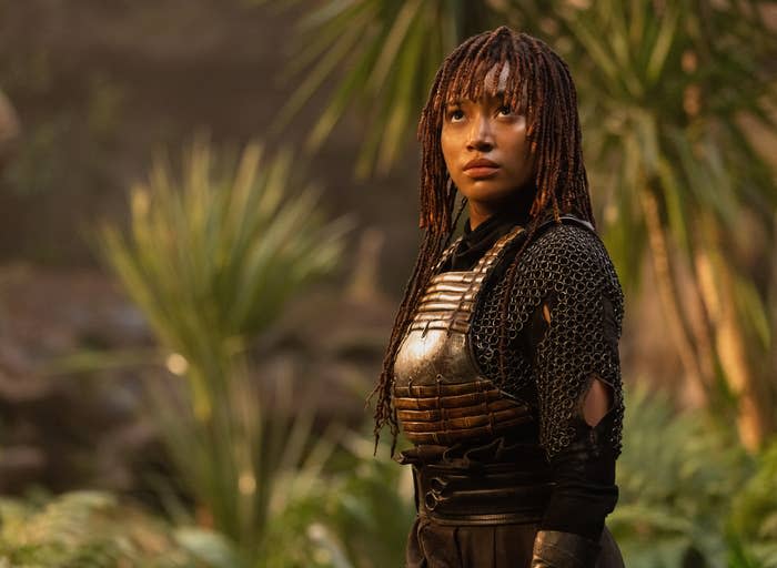 Amandla Stenberg in chainmail armor and leather chest piece, with braided hair, stands in a lush, green jungle setting