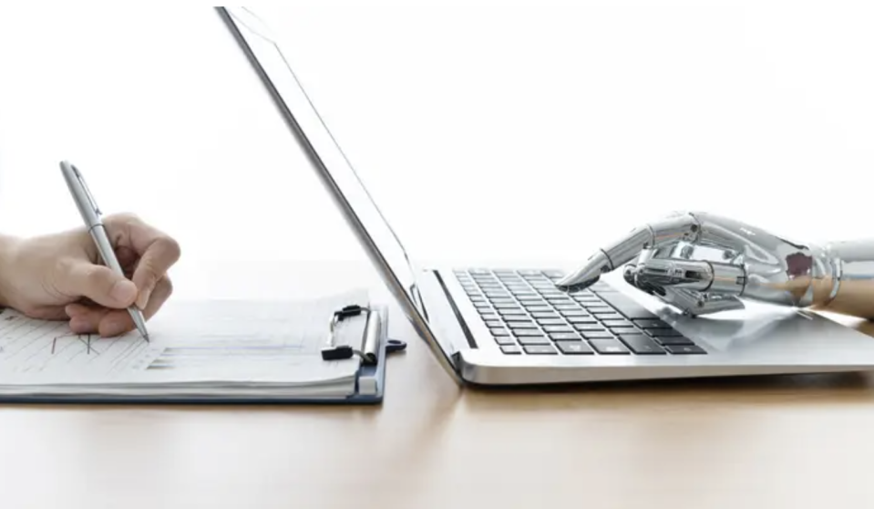 a human hand writing on a notepad next to a robot hand typing on a laptop