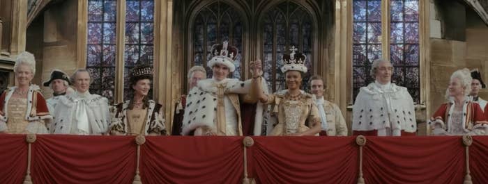 In a scene from 'Queen Charlotte: A Bridgerton Story" King George III and Queen Charlotte hold their raised hands on a castle balcony surrounded by dignitaries