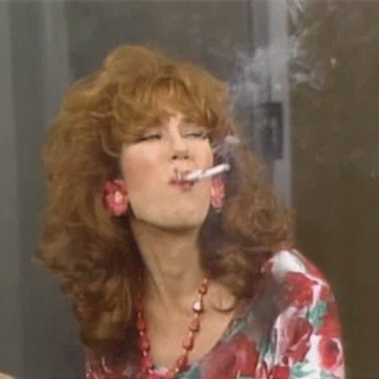 A woman with curly hair and dressed in a floral outfit smokes two cigarettes simultaneously, with smoke surrounding her face