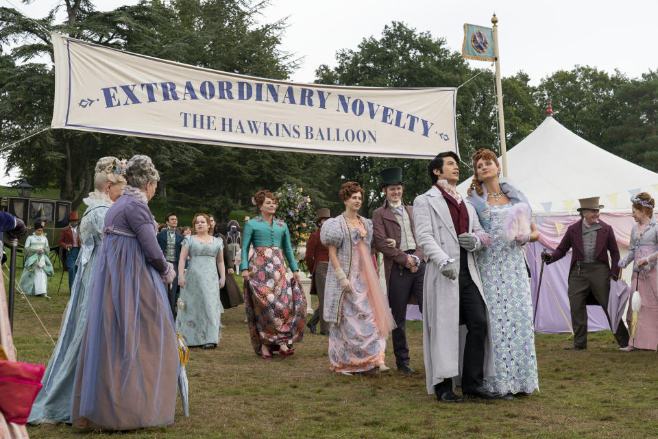 A group of people in colorful Regency attire at a fair under a banner that reads "Extraordinary Novelty: The Hawkins Balloon"