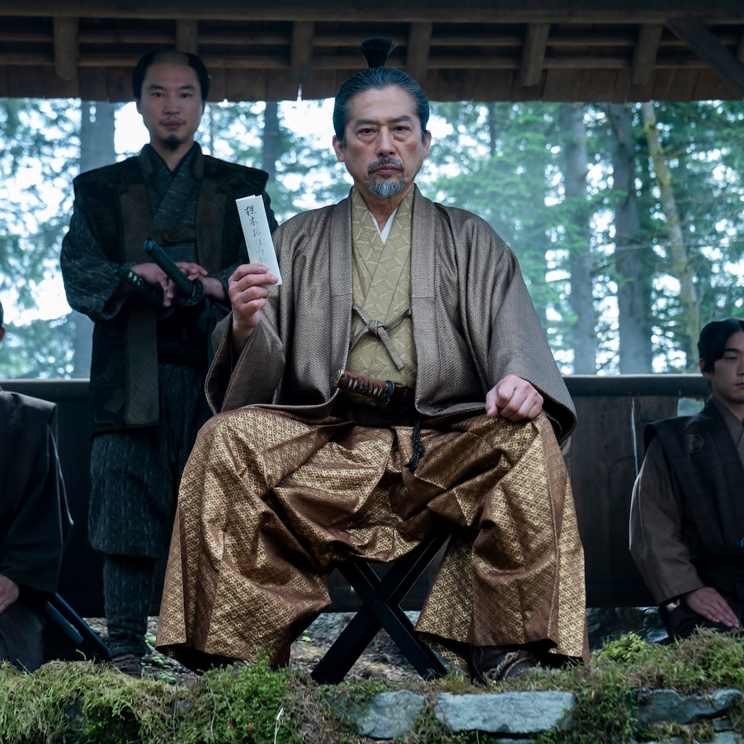  A man in gold japanese period wear (Hiroyuki Sanada as Yoshii Toranaga) sits while holding up a white letter, with one man standing behind him and two more kneeling at each side, in 'shogun' (2024). 