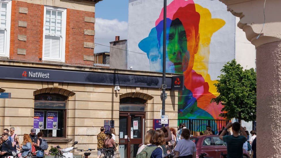 A colourful 3D like piece of street art picturing a man wearing a hat. Crowds of people are filling the street