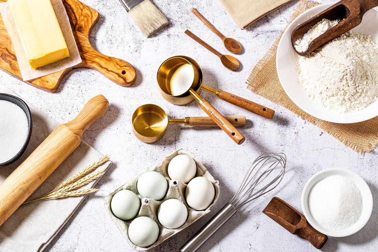 Top view of various bakery ingredients and items such as eggs, butter, flour, sugar, rolling pin, measure cups, hand whisk and serving scoop disposed all around the image on a bakery concept background