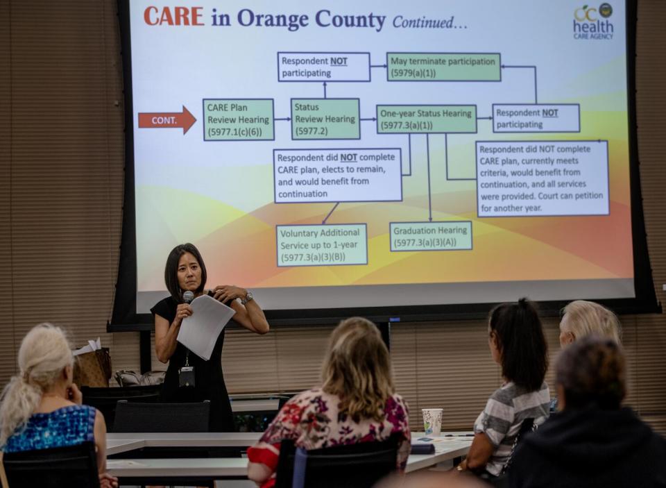 A woman holds a mic and documents stands in front of a projector screen facing an audience