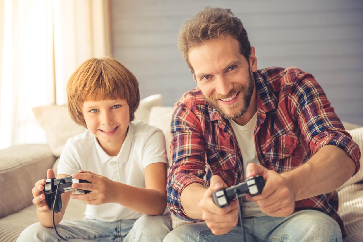 Cute boy and his handsome father are playing game console, looking at camera and smiling while sitting on couch at home