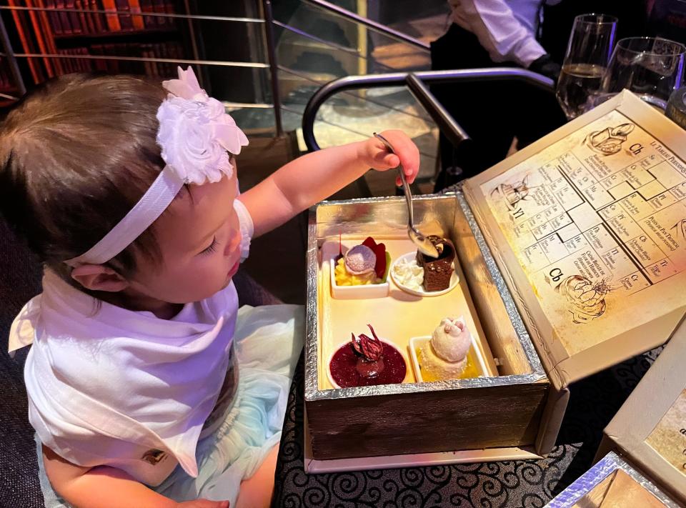 A toddler in a white headband with floral detail, blue skirt, and white shirt and bib holds a spoon and digs into a piece of cake. A treasure chest-like dish containing a variety of desserts sits in front of her