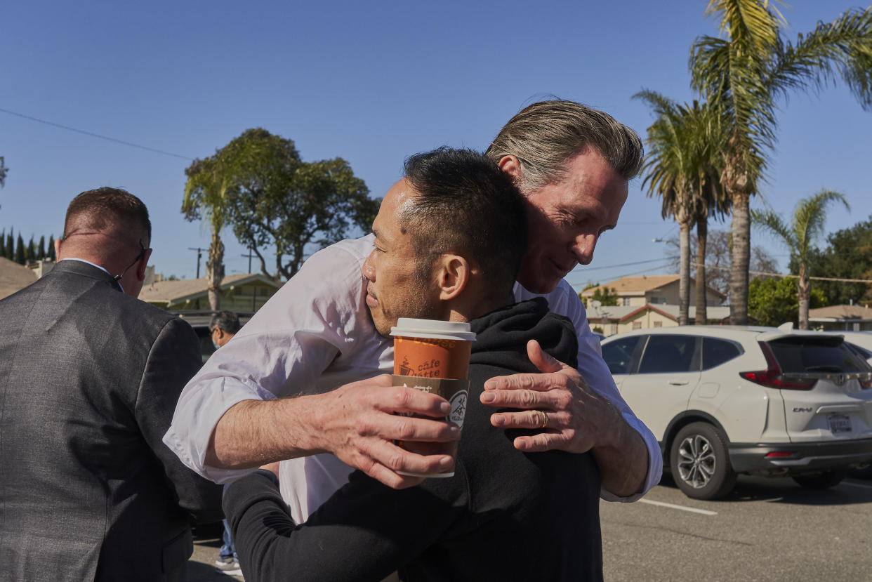 Gov. Gavin Newsom, holding a portable cup of coffee, hugs Thinh Luong in a parking lot, against a backdrop of palm trees.