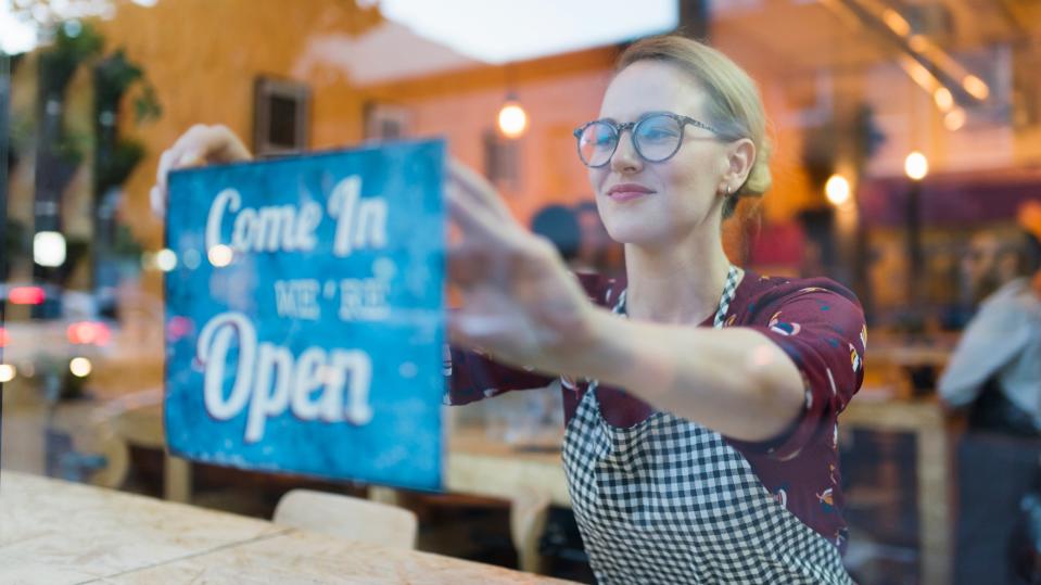 A woman putting up a "come in, we're open" sign on the window of her business.