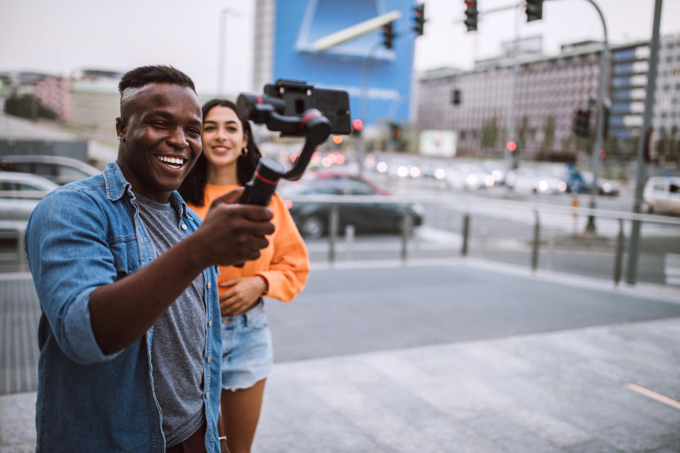 A man and woman smile while recording on a smartphone mounted on a gimbal, standing at a busy city intersection