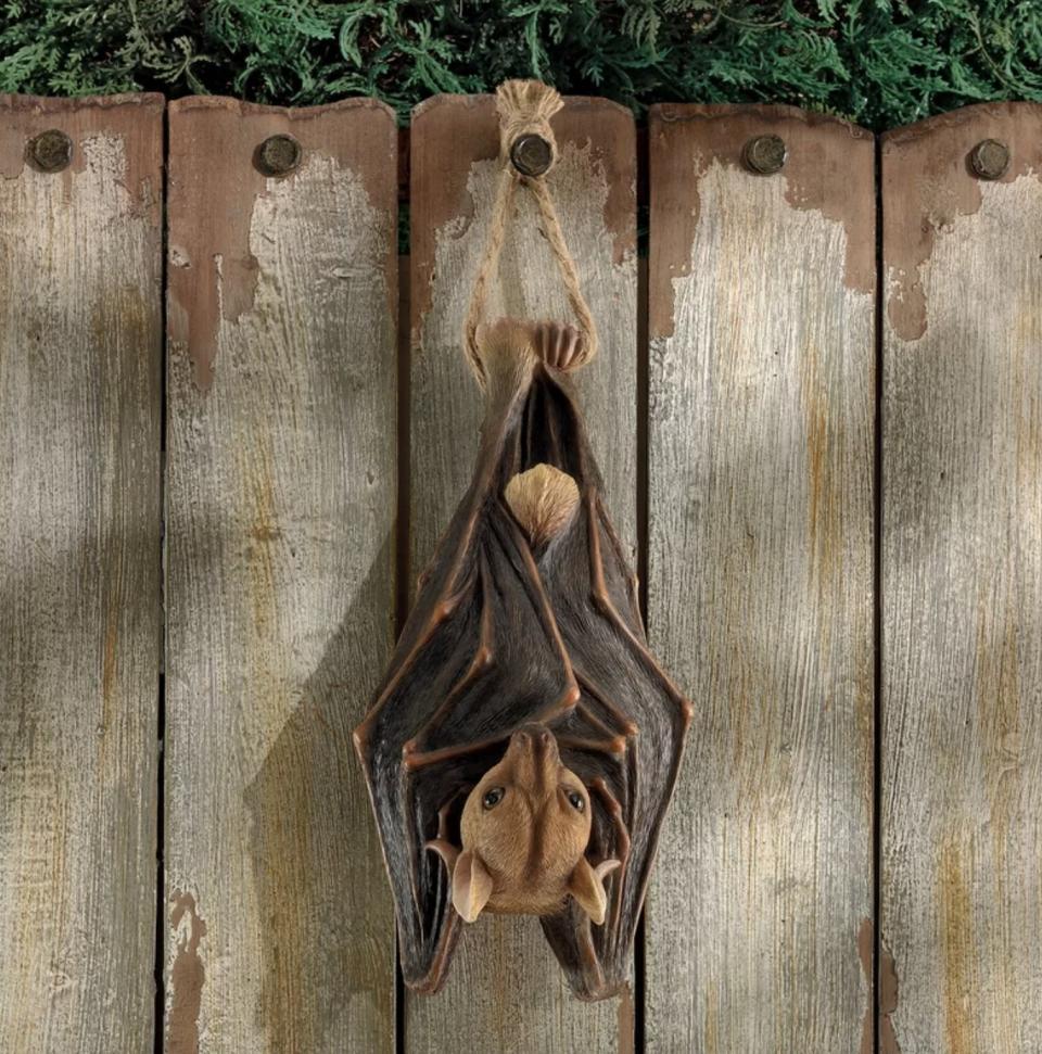 The brown and tan bat is hanging from a fence with its wings wrapped around itself and it's face peeking out