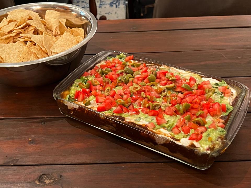 Finished 7-layer dip in a clear casserole dish and sour cream, guacamole, tomatoes, and olives on top. A bowl of tortilla chips sits beside the dish