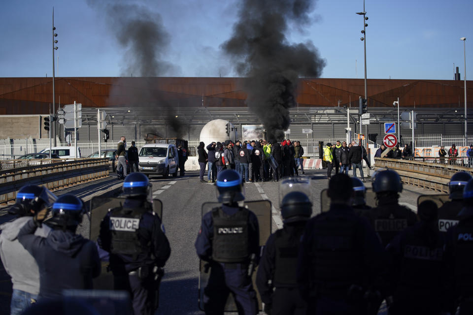 Dock workers face riot police as they stand in front of a burning barricade next the port of Marseille southern France, Wednesday, March 22, 2023. The bill pushed through by President Emmanuel Macron without lawmakers' approval still faces a review by the Constitutional Council before it can be signed into law. Meanwhile, oil shipments in the country were disrupted amid strikes at several refineries in western and southern France. (AP Photo/Daniel Cole)