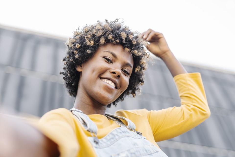 carefree afro woman with headphones looking at camera and smiling while taking a selfie outdoors