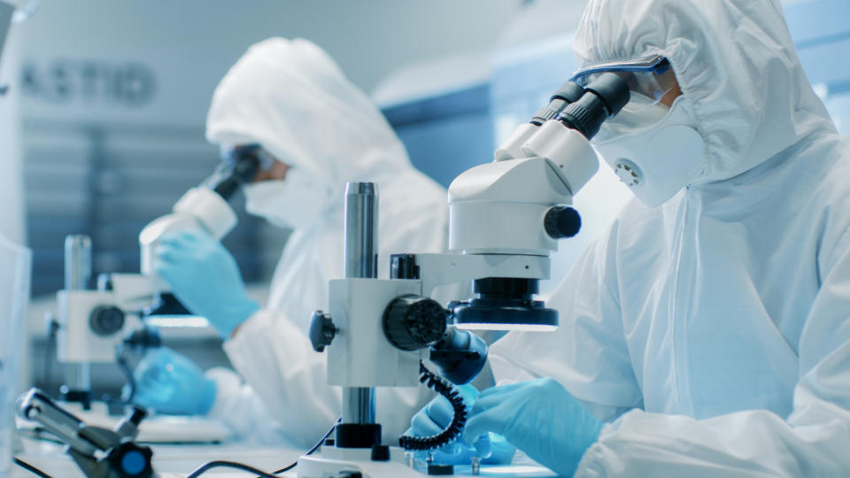 Two people in full-coverage lab suits looking through microscopes.