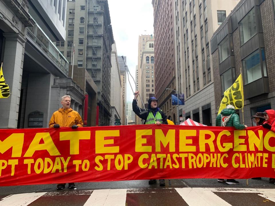 four people hold a red and yellow banner across a city street one person waves a crutch in the air