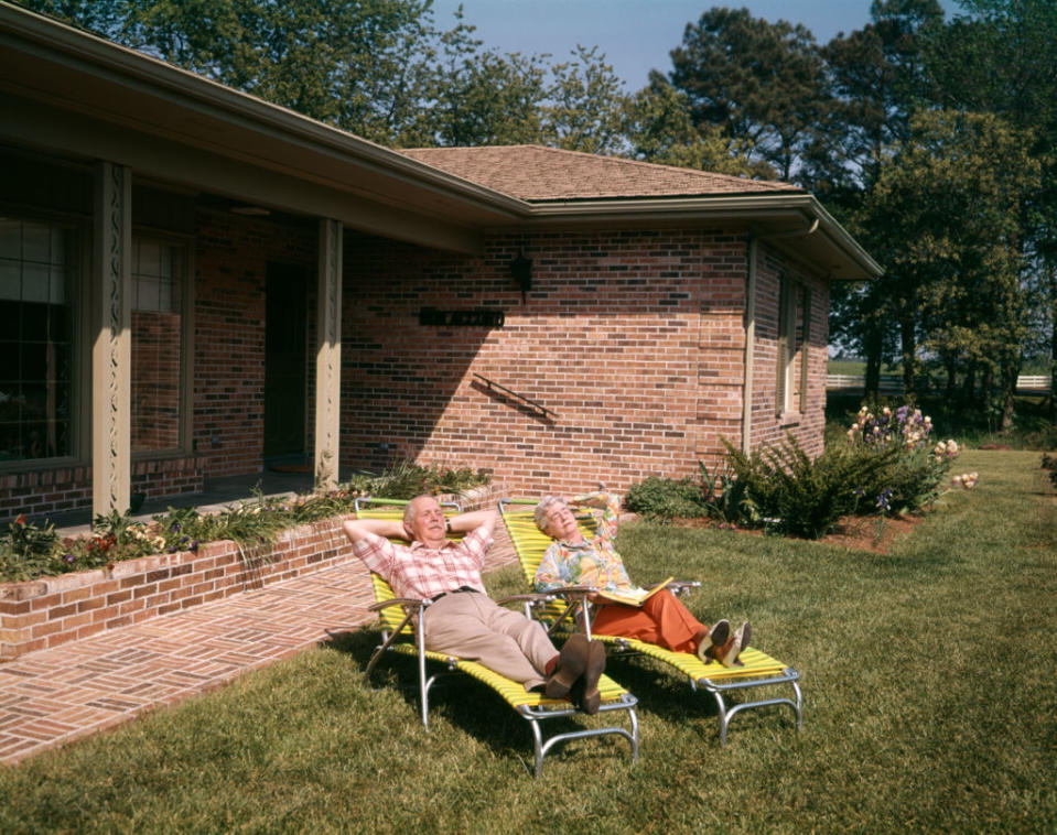 An elderly couple relaxes on lounge chairs in their yard outside a brick house. The man is reclining with his arms behind his head, while the woman reads a book