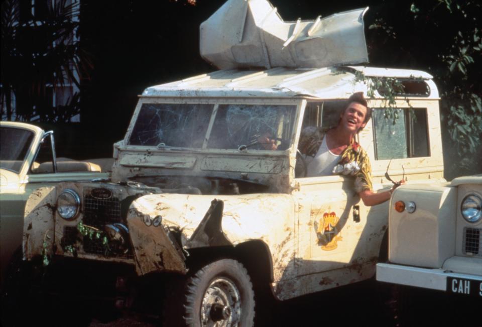 Jim Carrey as Ace Ventura sitting inside a Jeep, hanging out the window smiling