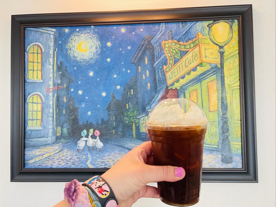 The writer holds cup of coffee to go with a night sky and city scene painting in background