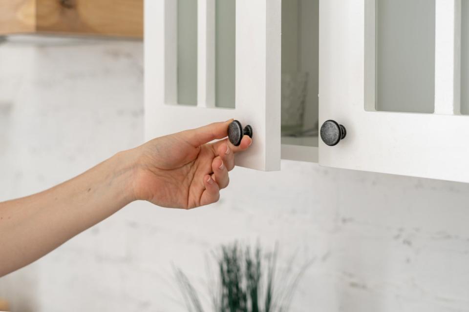 A woman's hand opening a white cabinet door by pulling on black metal knob.